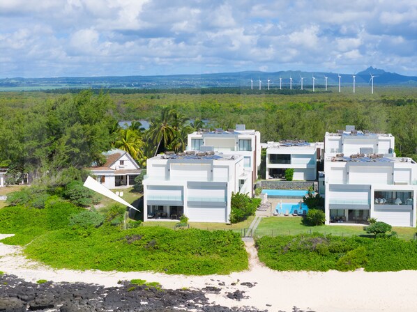 A beachfront property with multiple modern buildings, a swimming pool, and wind turbines in the background.