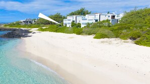 Um imóvel à beira-mar com uma praia de areia, águas turquesa cristalinas e um edifício moderno com vários andares e varandas.
