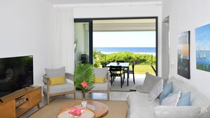 Sala de estar moderna com televisão, sofá e mesa de centro. Vista para o oceano através de uma porta de vidro.