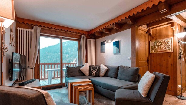 Un accogliente soggiorno con divano, poltrone e televisore. Soffitto in legno, finestra con tende e vista sull'esterno.