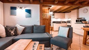 Un soggiorno accogliente con un divano componibile, un tavolino in legno e una poltrona. Si intravede sullo sfondo una cucina con mobili in legno e un'area pranzo con tavolo e sedie in legno.