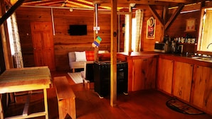 Een rustiek houten interieur met een eettafel, een bank, een keukenhoek met fornuis en gootsteen, en een aan de muur bevestigde televisie.