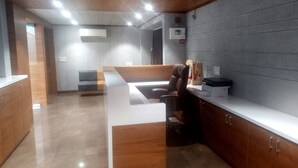 A reception area with a wooden reception desk, a seating area with a sofa, and a grey wall with a fire alarm. - Hotel Prime (Gandhinagar)
