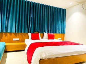 A hotel room with a wooden headboard, a large bed with red and white bedding, and a blue curtain. - Hotel Prime (Gandhinagar)
