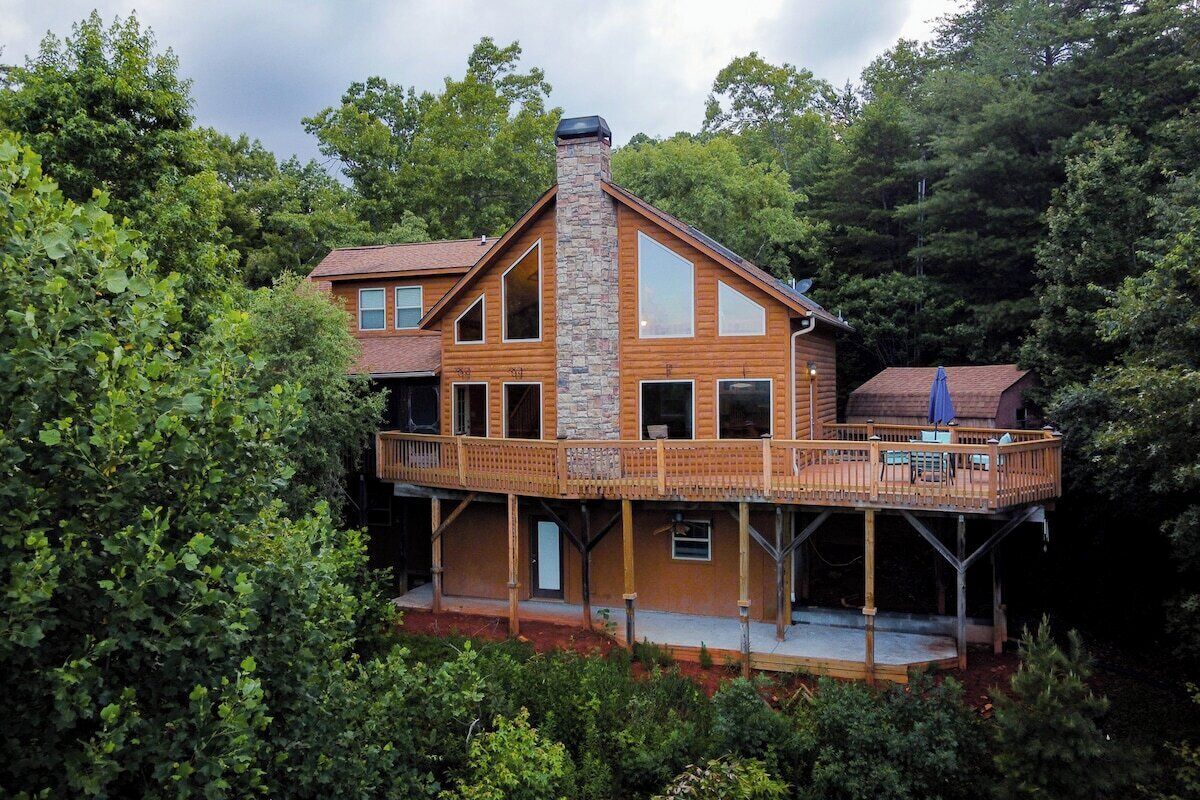 A large log cabin with a stone chimney, multiple windows, and a deck with outdoor furniture.