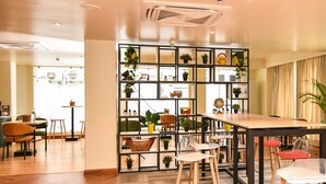Un interior moderno con suelo de madera, una mesa central con taburetes, una estantería decorativa con plantas y vistas a la calle.