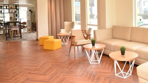 Un moderno vestíbulo de hotel con piso de madera en patrón de espiga, un sofá, sillones, una mesita redonda y un ventanal con vistas a la ciudad.