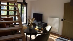 A modern living room with a wooden ladder, a dining table with chairs, a TV, a fireplace, and a large window with a view of trees. - Guesthouse with jacuzzi in picturesque Leiedorp (Zulte)