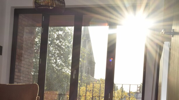 A room with a large window, a wooden floor, a brown chair, a small table, and a hanging light fixture. - Guesthouse with jacuzzi in picturesque Leiedorp (Zulte)