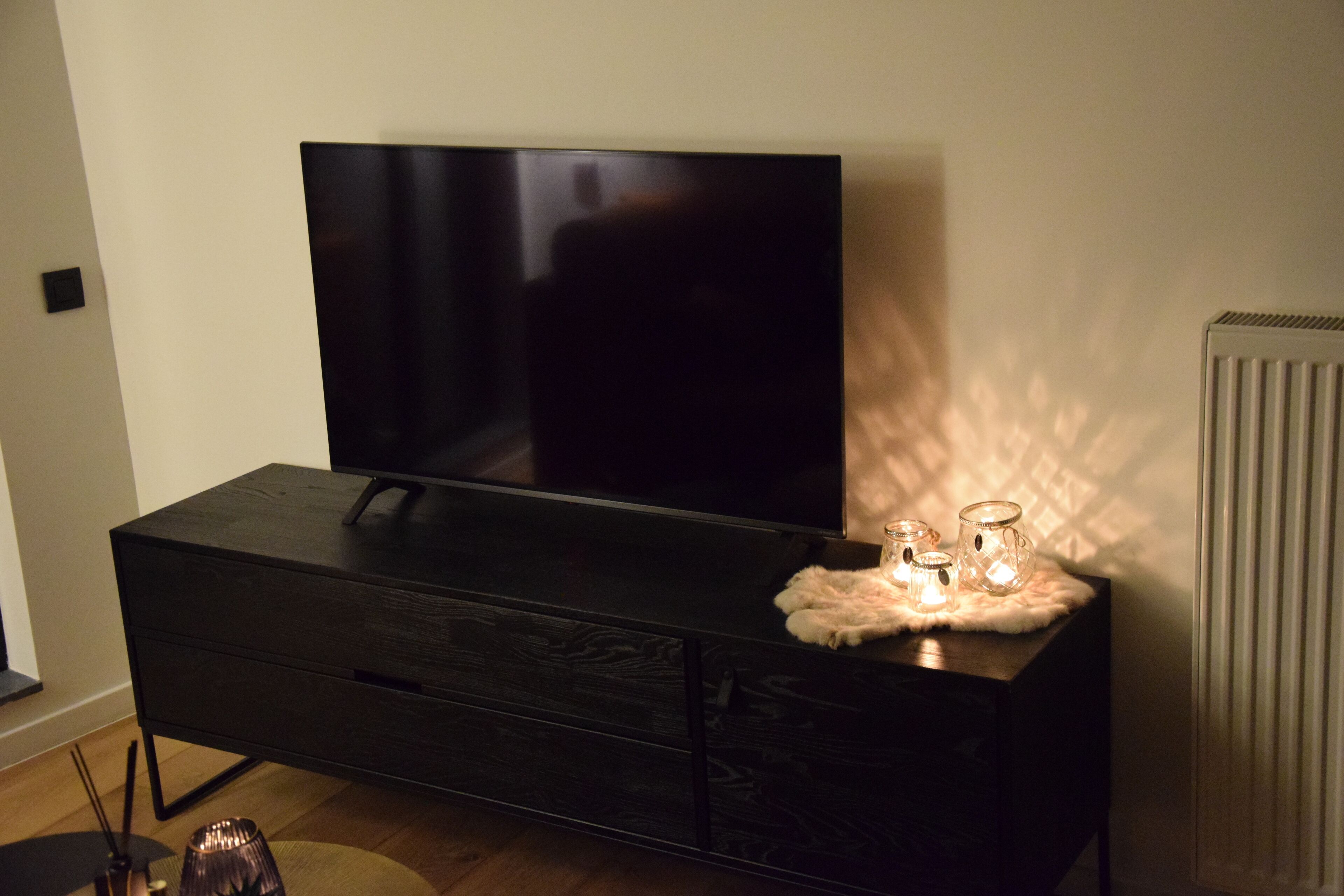 Ein modernes Wohnzimmer mit einem großen Flachbildfernseher auf einem dunklen Holzschrank, einem kleinen Tisch mit Kerzen und einer Heizung an der Wand.