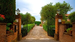 A cobblestone pathway leading to a gated entrance with brick walls and metal gates, flanked by greenery and potted plants.