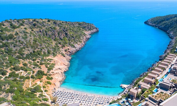 Vue aérienne d’une station balnéaire avec une plage, un terrain rocheux et une mer d’un bleu limpide.