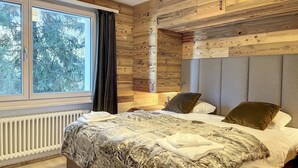 A bedroom with a large window, wooden panel walls, a bed with multiple pillows, and a radiator. - Living room with eating area, separate kitchen with dishwasher, one bedroom with 2 single beds, one (Verbier)