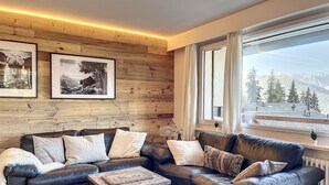 A modern living room with a wooden wall, a black leather sofa, and a view of mountains through large windows. - Living room with eating area, separate kitchen with dishwasher, one bedroom with 2 single beds, one (Verbier)