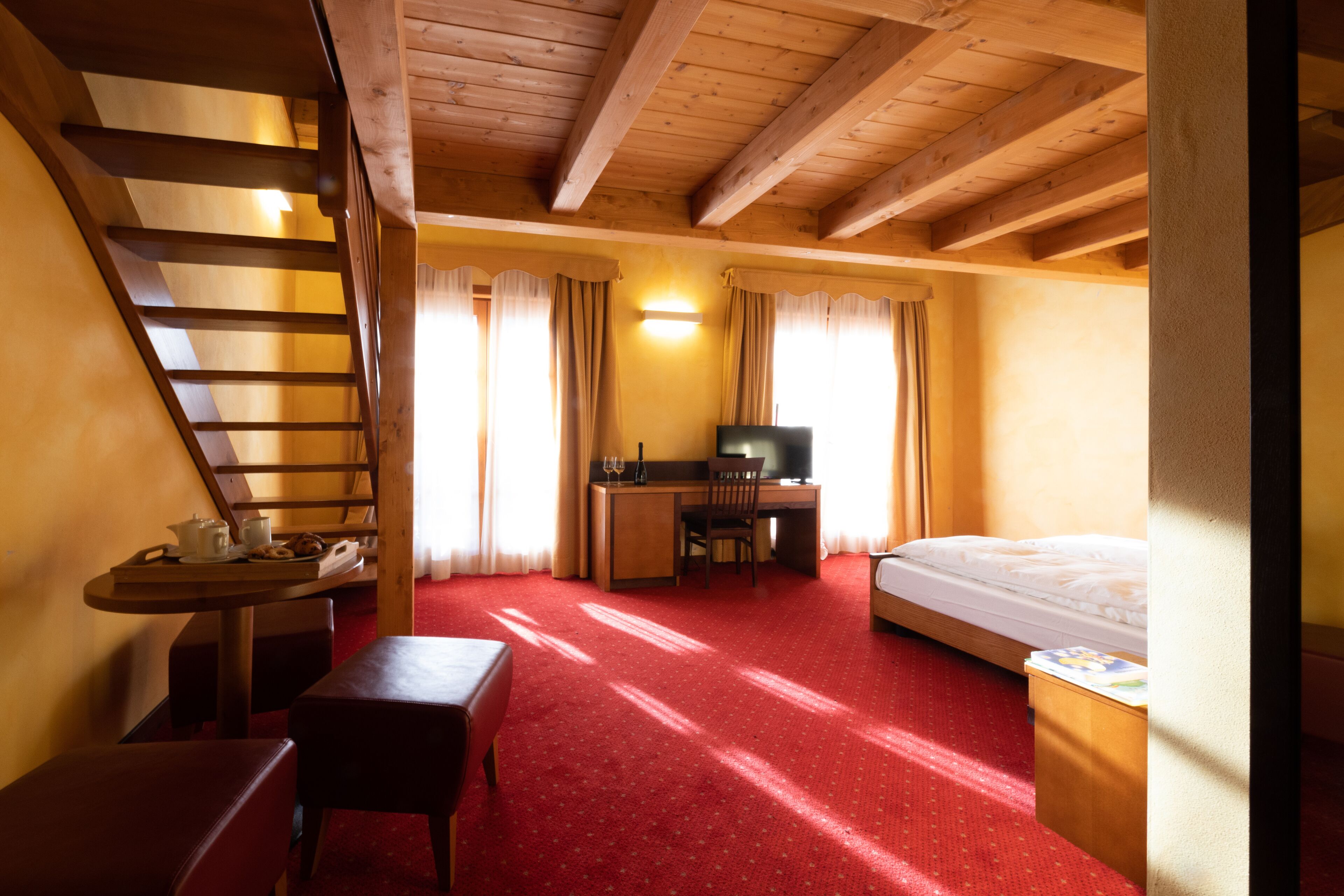 Habitación de hotel con techo de madera, una cama, un escritorio con silla, una mesita con lámpara y una escalera.