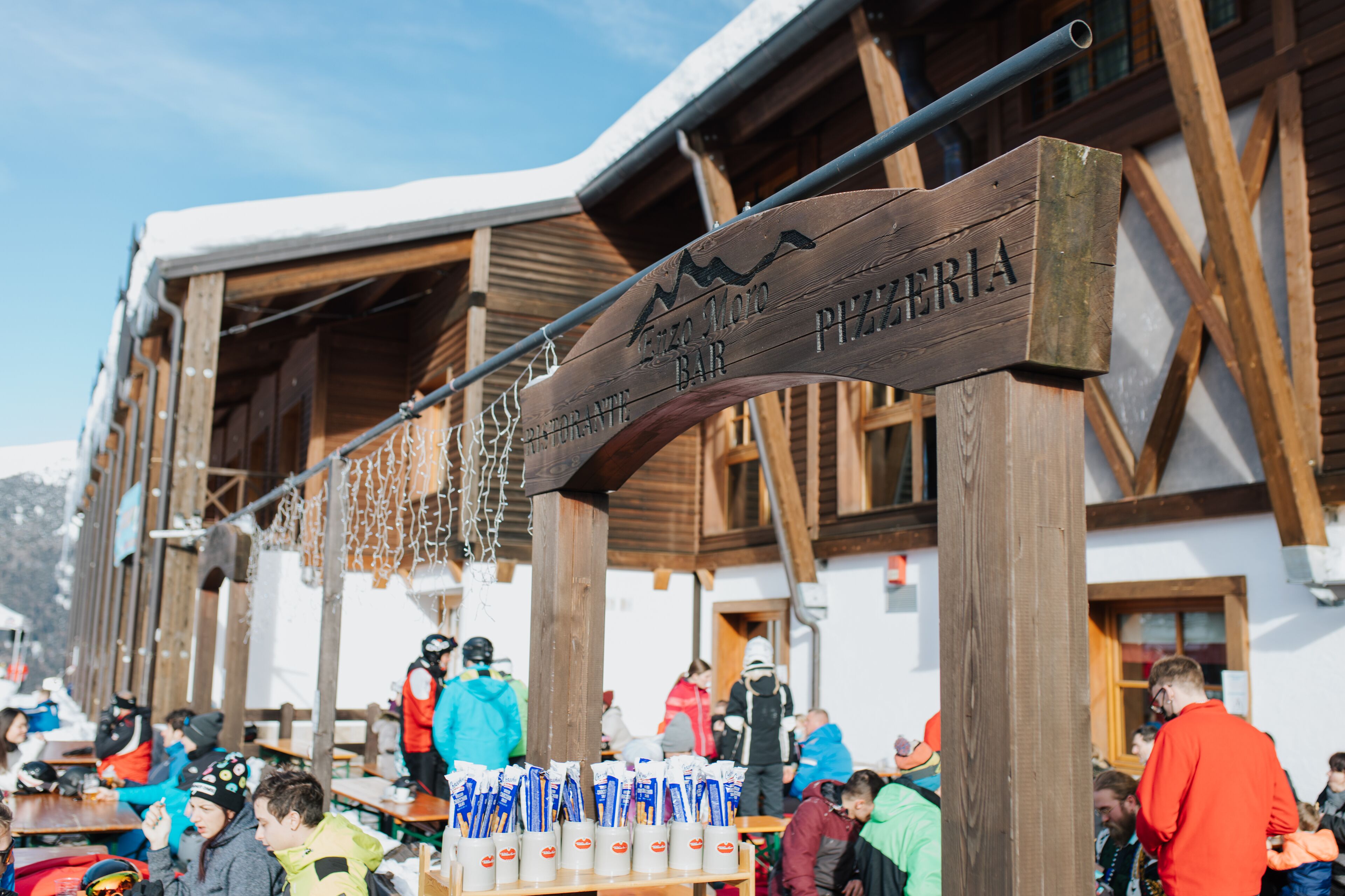 A wooden sign for a bar with a mountain peak logo, wooden beams, and people in winter clothing.