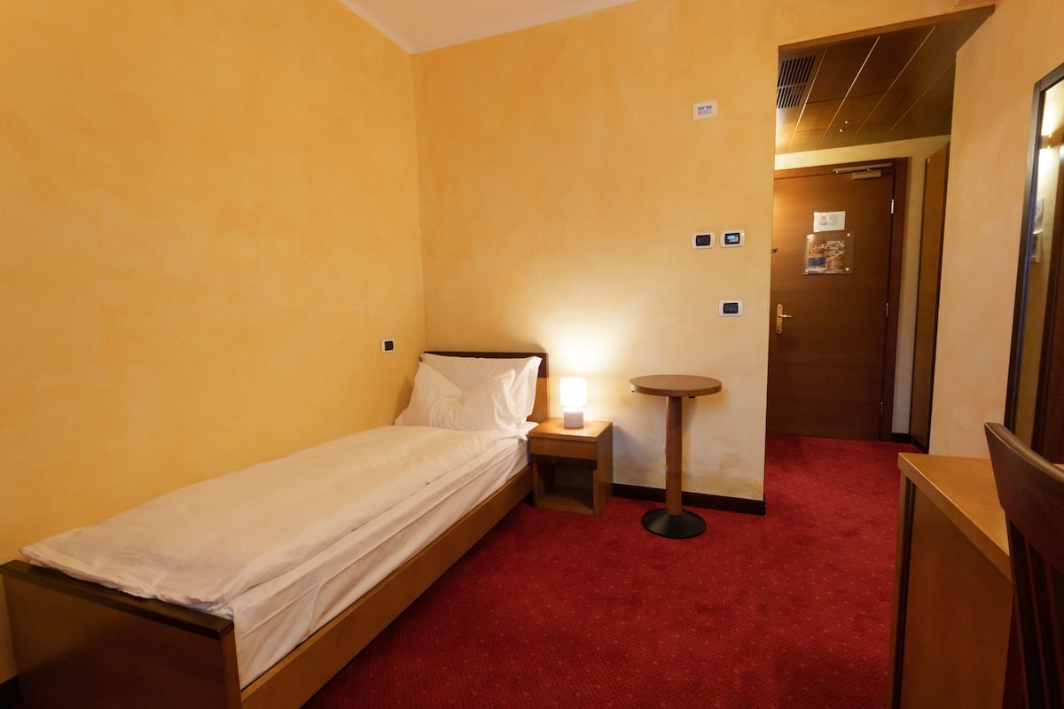 Ein Hotelzimmer mit einem Einzelbett, einem Nachttisch mit Lampe, einem kleinen runden Tisch und einem roten Teppichboden.