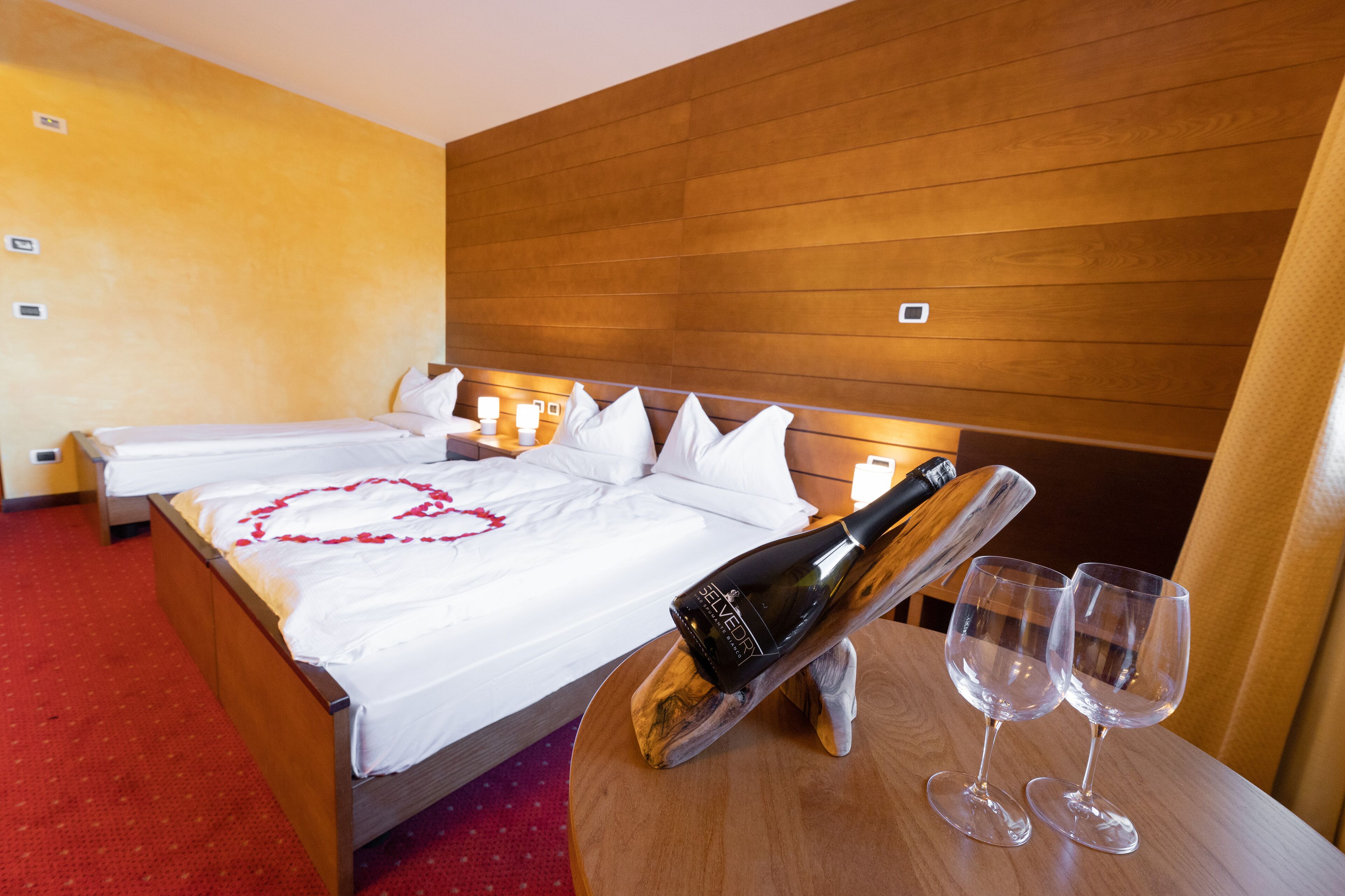 Ein Hotelzimmer mit einem hölzernen Kopfteil, einem Bett mit weißen Leinen, einer herzförmigen Dekoration auf dem Bett, einer Champagnerflasche und zwei leere Weingläser auf einem Holztisch.