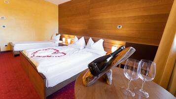Ein Hotelzimmer mit einem hölzernen Kopfteil, einem Bett mit weißen Leinen, einer herzförmigen Dekoration auf dem Bett, einer Champagnerflasche und zwei leere Weingläser auf einem Holztisch.