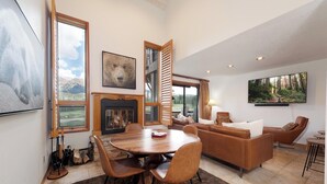 A spacious living room with a fireplace, a dining area with a wooden table and chairs, and a large window offering a mountain view. - Mid Century Modern Condo - Deck with Great Views - Heated Pool (Durango)