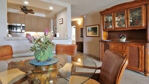 Un comedor con mesa de vidrio, sillas de mimbre, un mueble de madera con puertas de cristal y un ventilador de techo.