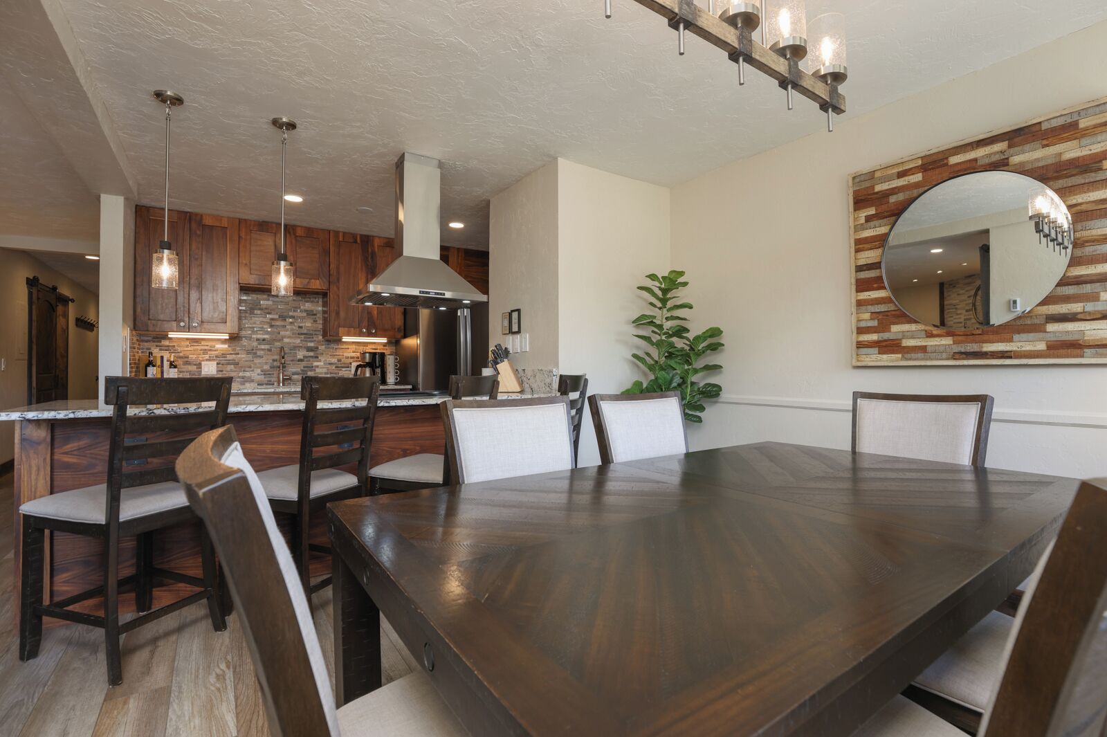 A modern dining area with a large wooden table and chairs, a kitchen with wooden cabinets and a brick backsplash, and a large mirror on the wall.