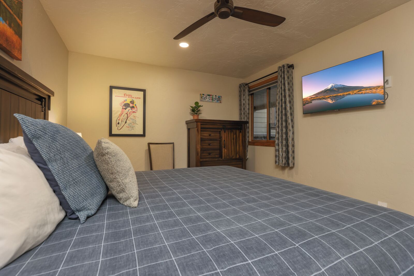 Une chambre à coucher avec un lit, des oreillers, un ventilateur de plafond, un tableau encadré, une fenêtre avec des rideaux et un téléviseur fixé au mur.