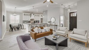 A modern living room with a brown sofa, a grey armchair, and a coffee table. There is a dining area with a table and chairs, and a kitchen in the background. - Trendy and Modern home w/ a Private Pool (Rowlett)