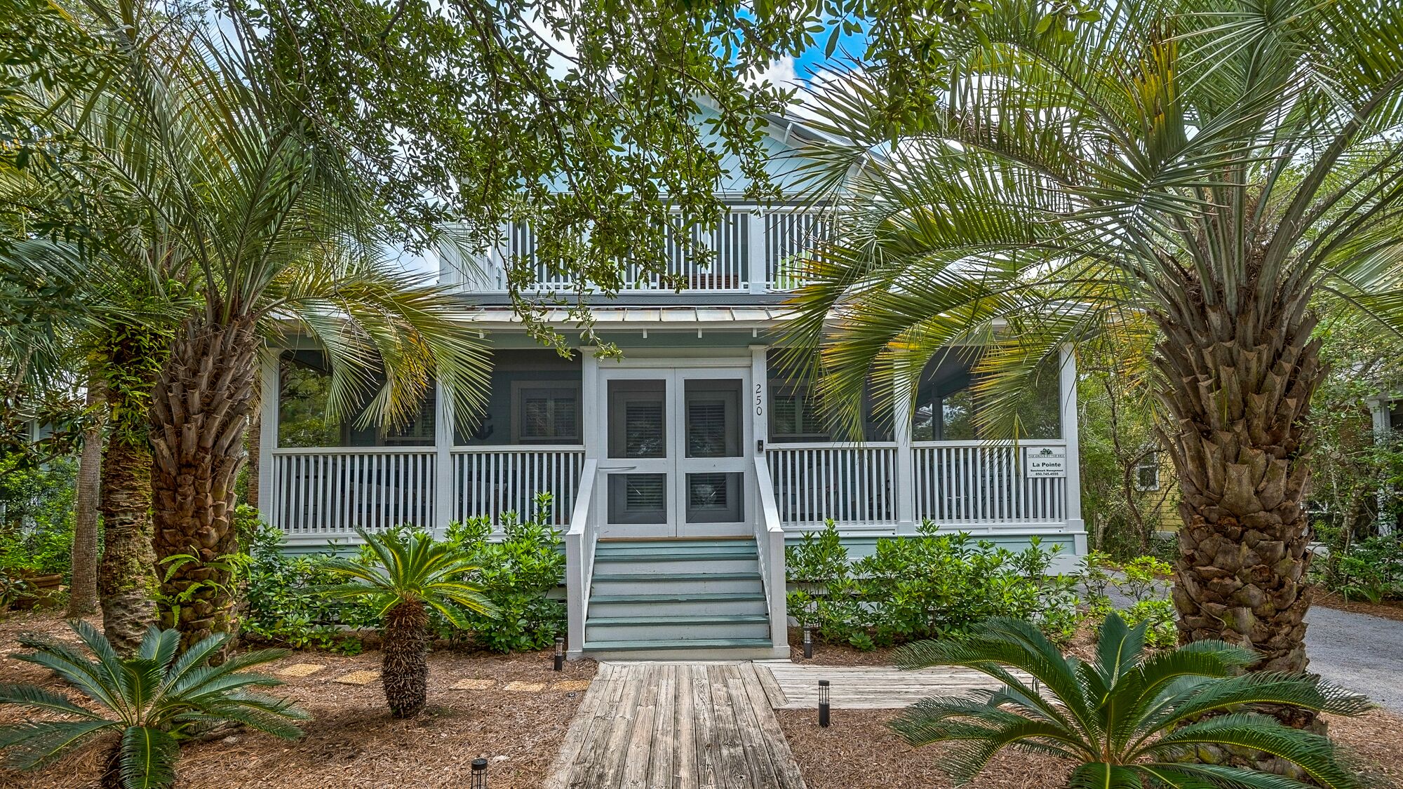 A two-story house with a porch, surrounded by tropical plants.