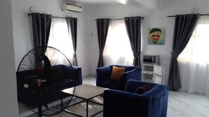 Una sala de estar con un sofá azul, una mesita, un ventilador de pie y un microondas.