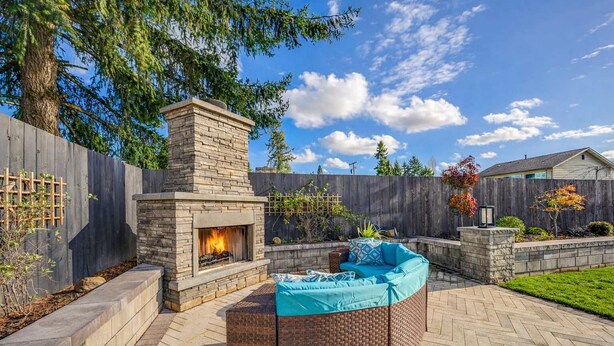 A stone fireplace with a fire, surrounded by a brick fire pit and a blue cushioned seating area. - Renton Oasis-Relaxing 3BR Home with Great Outdoor (Renton)