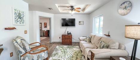 Un salon avec un ventilateur de plafond, un téléviseur, un canapé, un fauteuil et une horloge au mur.