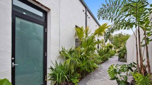 A narrow pathway flanked by lush greenery and a white building with a black-framed door. - Claudia | Sense of Calm and Relaxation,  Walking Distance to the Excitement - PO (New Orleans)