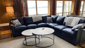 A modern living room with a dark leather sectional sofa, two round coffee tables, a lamp, and a view of a lake through large windows. - Lake & Mountain Retreat - Summer Adventures & Winter Escape (Center Ossipee)