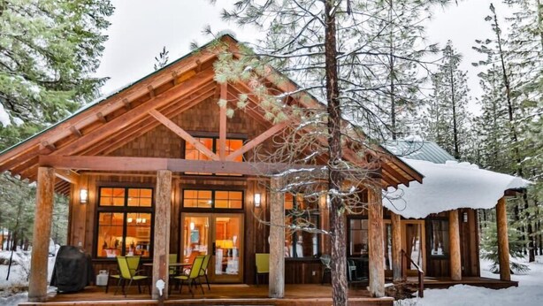 En stuga byggd av trä med en inglasad veranda och en matplats, omgiven av snö och träd.