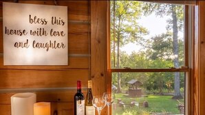 A wooden cabin interior with a sign reading "bless this house with love and laughter," a candle, two wine bottles, and glasses on a wooden table by a window overlooking a garden.