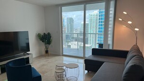 A modern living room with a large TV, a sofa, a blue chair, a small round table, and a potted plant. - *New* Elegant Apt Brickell 5 min. From Beach (Miami)