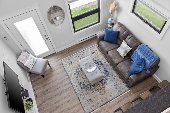 A modern living room with a brown leather sofa, a coffee table, and a patterned rug. There is a television on a stand, a small side table with a vase, and a window with a view of the outdoors.