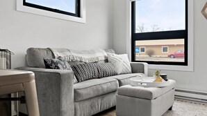 A modern living room with a grey sofa, a matching ottoman, and a large window offering a view of a building and a red car outside. - TC Loft Near Downtown w/ Open Layout (Traverse City)