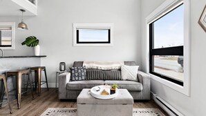 A modern living room with a grey sofa, a round ottoman, a small table, and a window with a view of the outside. - TC Loft Near Downtown w/ Open Layout (Traverse City)