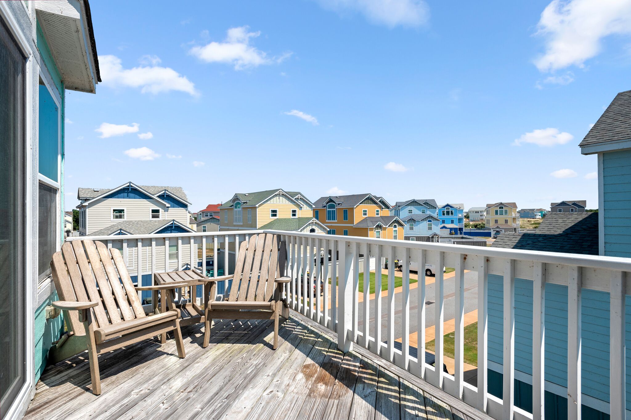 Une terrasse en bois avec deux chaises Adirondack et une balustrade blanche, d’où l’on peut admirer une rangée de maisons aux couleurs vives.