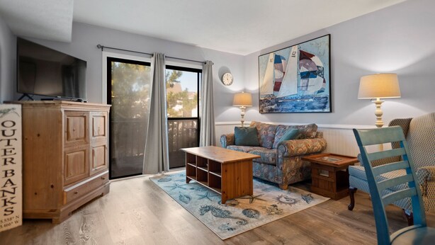 A cozy living room with a wooden cabinet, a sofa, a coffee table, and a rug with a nautical theme. - Beach Place: Oceanside | Short Walk to the Beach (Corolla)