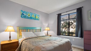 A bedroom with a bed, two bedside tables with lamps, a window with curtains, and a painting on the wall. - Beach Place: Oceanside | Short Walk to the Beach (Corolla)