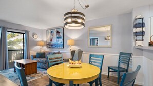 A dining area with a round yellow table, blue chairs, and a chandelier. - Beach Place: Oceanside | Short Walk to the Beach (Corolla)