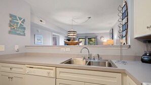 A kitchen with a double sink, white cabinets, a dishwasher, and a hanging light fixture. - Beach Place: Oceanside | Short Walk to the Beach (Corolla)
