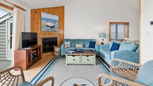 A living room with a blue sofa, a wooden entertainment unit, a fireplace, and a large painting of a boat on the wall. - SeaFinn: Semi-Oceanfront | Pool Access | Dogs OK (Nags Head)