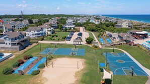 An aerial view of a coastal residential area with multiple houses, a beach, and a sports court complex. - Plum Paws: Pool | Hot Tub | Sound Views | Dogs OK (Corolla)