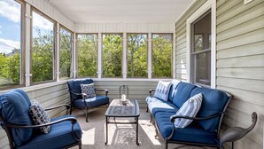 Un porche cubierto con muebles azules y una mesa, ventanas amplias y vistas a árboles.
