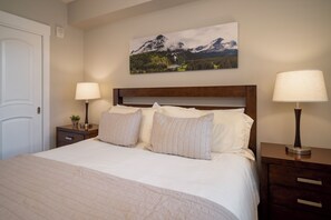 A neatly made bed with a wooden headboard, two bedside tables with lamps, a wall-mounted painting of a mountain landscape, and a door to the left. - Fraser Crossing 1 Bedroom modern and comfortable (Winter Park)
