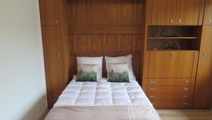 Een bed met een gewatteerde witte dekbedovertrek en twee kussens met bosmotieven, voor een houten garderobe met meerdere lades en planken.